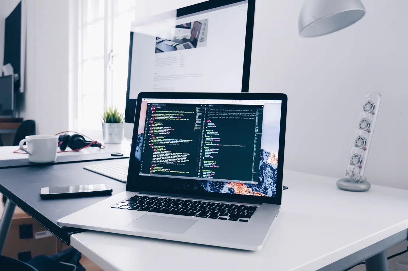 Laptop on a desk showing website code, with a second monitor behind it